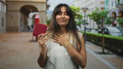Young latina brunette woman holding small red wallet with one hand, other hand on chest, standing...