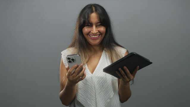Young brunette woman holding smartphone and tablet in grey studio, smiling with head tilted up and laughing while checking devices; joy connection.