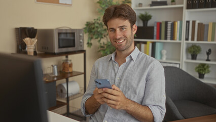Young man holding smartphone and smiling while seated at desk in office building; calm productivity.