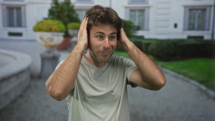 Man with hands to head, mouth open and raised eyebrows, standing near a white building entrance and trimmed hedges in an outdoor courtyard; surprise curiosity amusement.