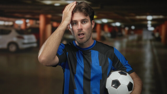 Man in blue striped soccer jersey holding a black and white ball, hand on forehead in parking building with blurred cars and concrete pillars; solo frustration. - Powered by Adobe