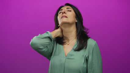 Hispanic woman grips neck to relieve muscle tension and stiffness against vibrant purple studio wall; discomfort.