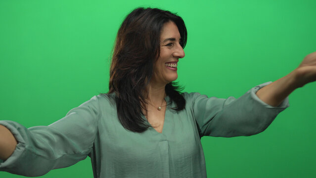 Woman in green blouse smiles while extending her arm and placing hand on chest against a bright green studio wall; joy.