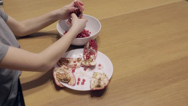 Two small hands split pomegranate segments and drop seeds into a bowl. Mid top view showing slow movements and tactile exploration.
