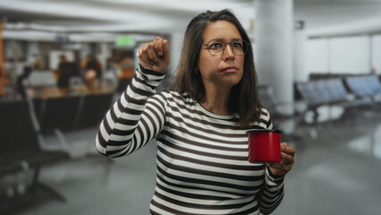 Brunette middle age woman holds red mug and raises left fist in crowded airport terminal seating...