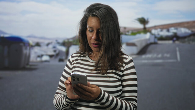 Middleaged hispanic woman tapping smartphone with hands, looking down on street near dumpster and white buildings; concentration connection. - Powered by Adobe