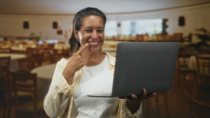 Woman hispanic middle age brunette smiling and pointing finger to cheek while holding laptop and looking at screen in building dining hall; joy connection.
