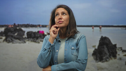 Woman touching temple with finger, other arm crossed and worried expression against studio beach backdrop; concern reflection.