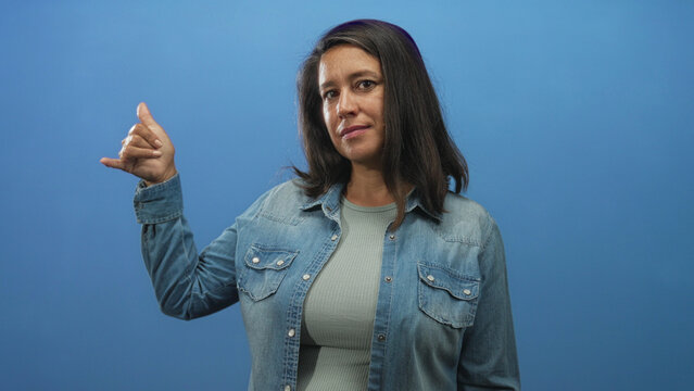 Woman middleaged hispanic brunette raises left hand with index and pinky extended in a horns hand sign against blue studio backdrop wearing denim jacket; confidence greeting.
