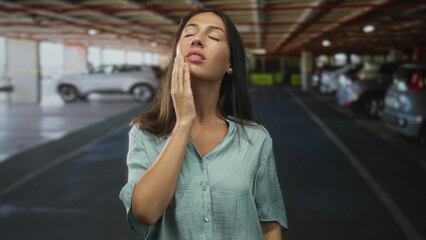 Fototapeta premium Young brunette woman touching her face with hand, covering mouth and rubbing cheek while standing in a building parking garage next to parked cars; fatigue inconvenience.