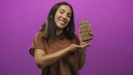 Hispanic woman pointing at a large chocolate bar with a presenting pose in a purple studio; playful indulgence.