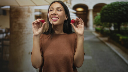Woman smiling and holding two strawberries between her fingers with hands raised on a cobblestone street; joyful summer picnic.