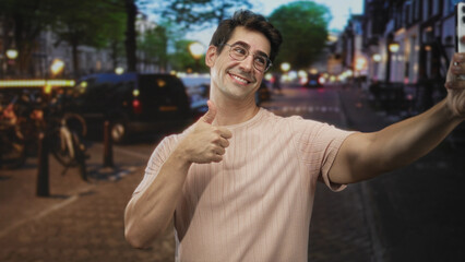 Fototapeta premium Man taking selfie with smartphone while giving thumbs up on a busy evening city street; playful confidence.