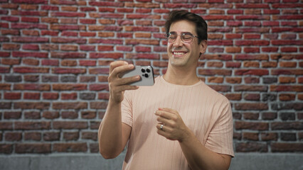 Man holding smartphone with visible hands, smiling while looking at the screen in a brick wall studio backdrop; joyful connection.