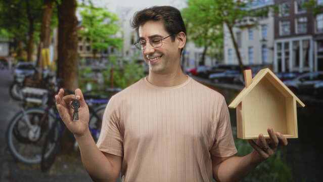 Man holding keys between fingers and a small wooden house model in one hand, smiling, posed on a city canal street with bicycles and townhouse buildings visible; homeownership pride trust.