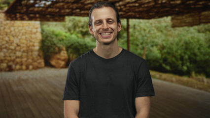 Man smiling and seated on a wooden deck in a forest pavilion under a woven canopy; serenity ease...