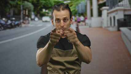 Man points both fingers directly at camera on street wearing apron and black t shirt, hands forward and visible; confidence invitation.
