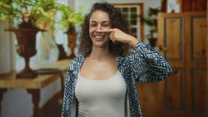Woman points finger to nose in a living room building wearing white tank top and patterned shirt; playful gesture.