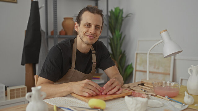 Man presses clay with hands on wooden board in studio, shaping a small ceramic bowl amid tools, sponge, and bowl of slip; calm creativity. - Powered by Adobe