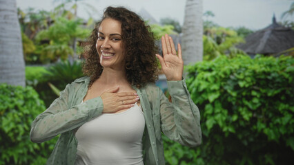 Woman with hand on chest and palm raised in forest park setting, wearing white tank top and sheer...