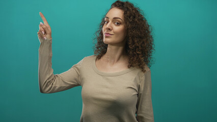 Fototapeta premium Young hispanic woman pointing finger with hand toward upper left while looking aside in studio with teal background; confidence focus.
