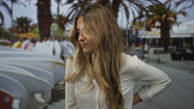 Woman winces with eyes closed and hand on hip by stacked surfboards in beachside surf shop; discomfort frustration anxiety unease. - Powered by Adobe