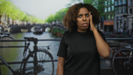 Young african american woman with hand on cheek showing toothache on a street beside an amsterdam canal and bridge, framed in outdoor urban scene; pain.