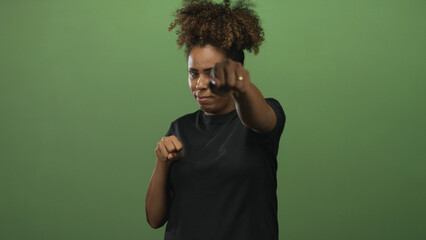 Young african american woman with clenched fists and raised hands in studio with green backdrop; determination strength focus.
