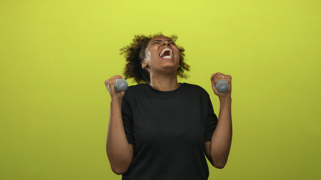 Young african american woman holding dumbbells and shouting in studio with vivid lime green backdrop; empowerment.