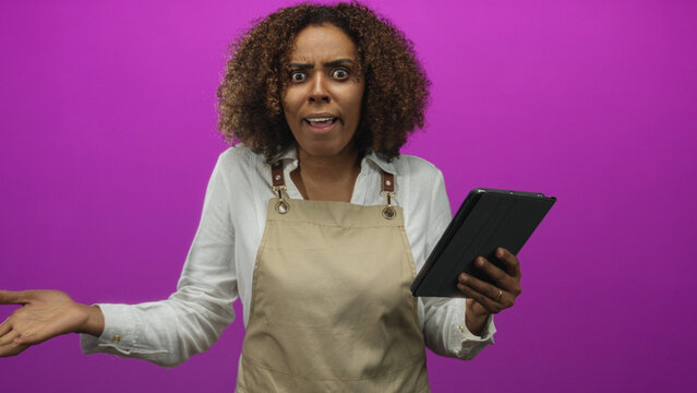 Young african american woman baker wearing apron holds tablet and makes fist gesture while checking orders in magenta studio; determination focus.
