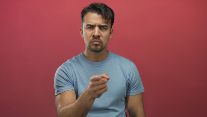 Young hispanic man in blue shirt pointing finger seriously against isolated red background showing...