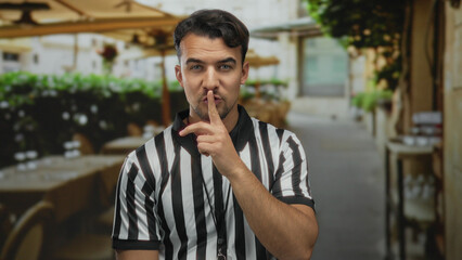 Young hispanic man in a referee shirt gestures silently on a terrace of an outdoor restaurant with tables and lush greenery creating a relaxed coffee shop vibe.