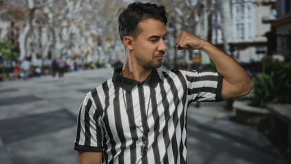 Young hispanic man in referee uniform flexing arm outdoors on city street showcasing determination and strength surrounded by urban environment and trees in background.