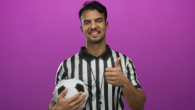 Young hispanic man wearing referee uniform holding soccer ball with thumbs up against isolated pink background, expressing positivity and sports enthusiasm.