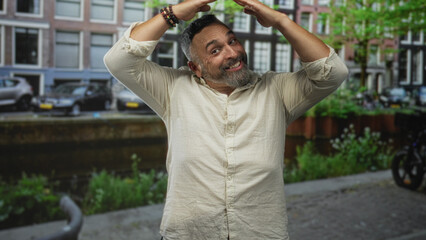 Man with beard smiling while hands form a roof over his head on street by a canal and parked cars, rolled sleeves visible, casual shirt; joy celebration.