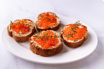 Fresh homemade canape made from full grain baguette, butter and salmon caviar deocrated with aromatic thyme twigs on white ceramic plate close up.