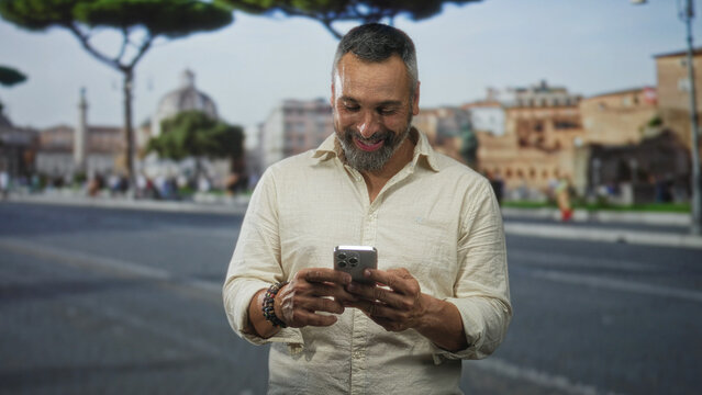 Middle aged man holding smartphone with hands visible and smiling on street by historic building; contentment connection.