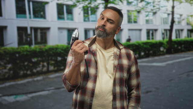 Man holding power drill in right hand on street, carpenter inspecting the tool with raised eyebrow and neutral pose; confidence experience.