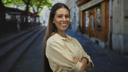 Fototapeta premium Woman smiling with arms crossed on street near cafe steps and wooden storefront; confidence approachability.