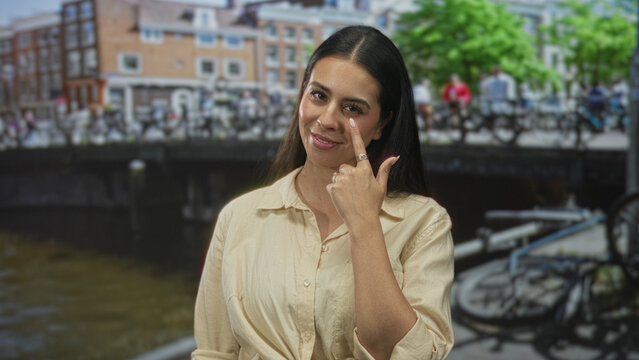 Woman points finger to eye on canal street in amsterdam, smiling with rings visible; playful confidence.