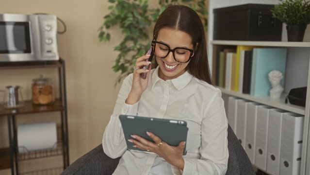 Woman holding tablet and phone to ear in office, smiling while seated in chair and wearing glasses; remote work productivity.
