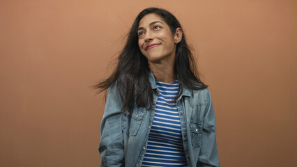 Woman smiling with hair blowing, wearing denim jacket and blue striped crop top in peach studio; contentment.