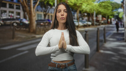 Young woman standing on street with hands forming a heart at her waist, belt and jeans visible; calm reflection.