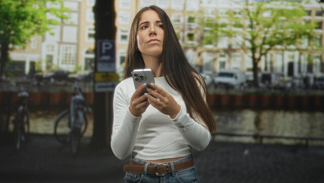 Woman in white top holding smartphone with hands tapping screen, long hair, standing by canal railing and bicycles on street; waiting concern. - Powered by Adobe