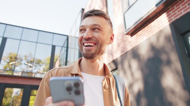 Attractive Caucasian male holding smartphone and smiling while enjoying lighthearted video content. Man carrying backpack standing near modern brick facade. Cheerful male taking brief pause.