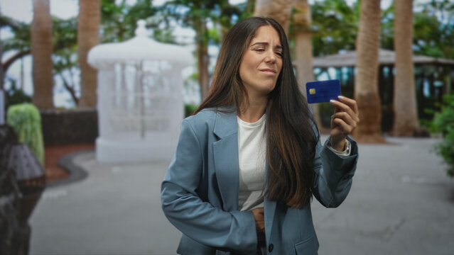 Young hispanic woman in blazer holding creditcard up on street, grimacing and clutching stomach; financial worry.