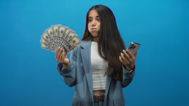 Woman fanning dollars and holding smartphone in studio while smiling at camera; financial success confident.