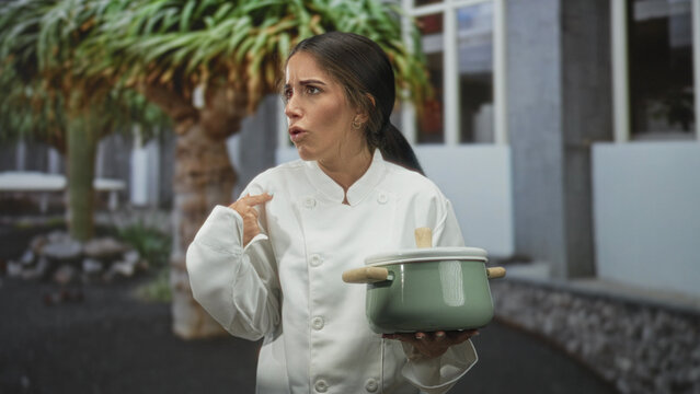 Woman chef in white coat holds green pot with wooden handles and points to self in courtyard; surprise uncertainty.