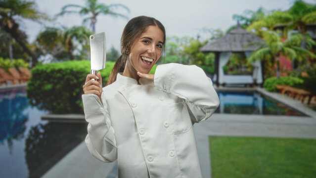 Young hispanic woman chef smiling and holding cleaver up in right hand beside a tropical pool gazebo building; confidence skill hospitality.