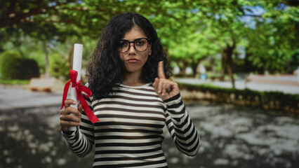 Woman holds rolled diploma with red ribbon and glasses, hand raised in forest pathway with trees...